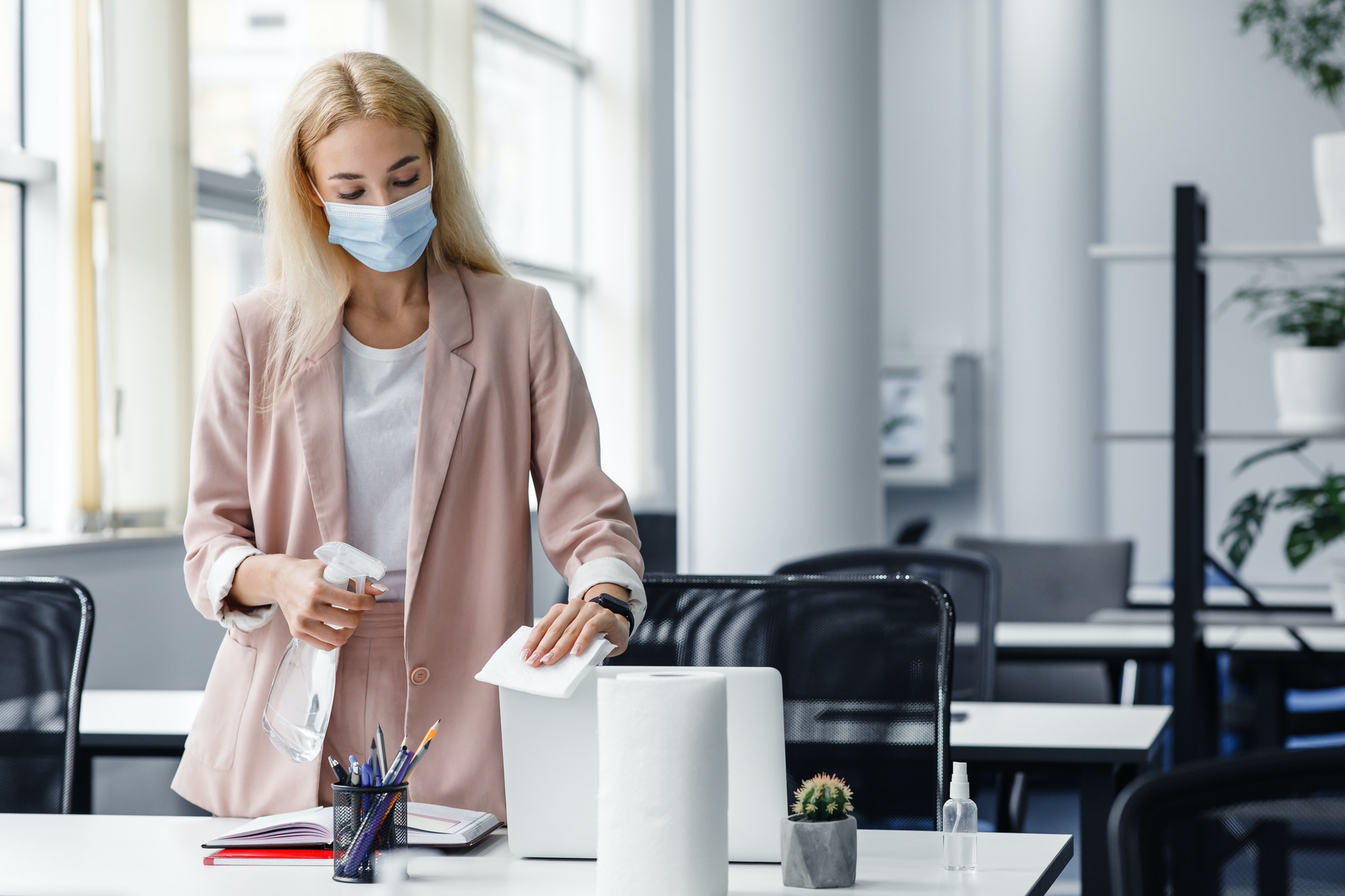 Hygiene in workplace in coworking office. Blonde business woman in suit and protective mask cleans